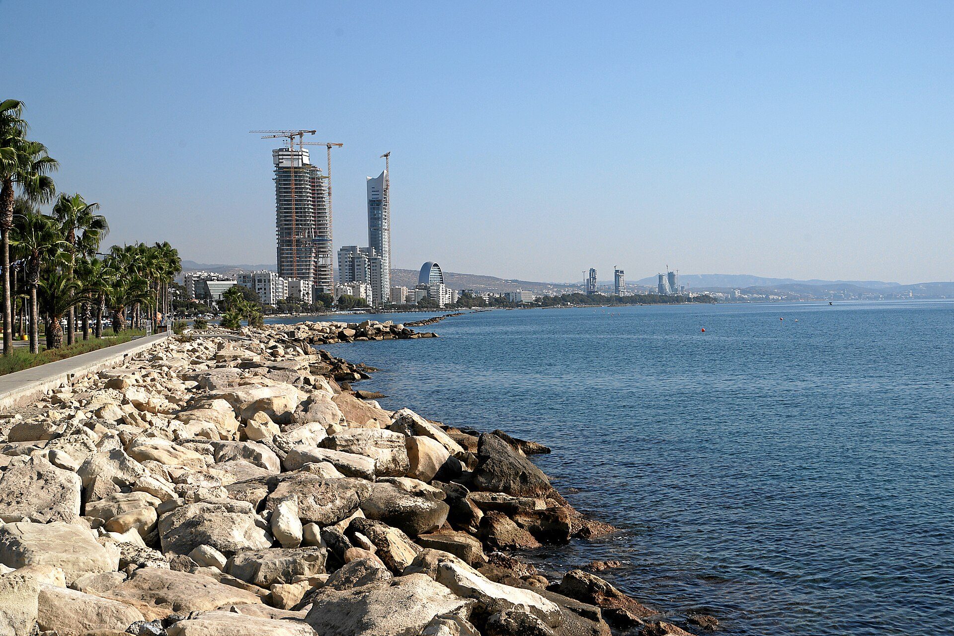 Βραχώδης ακτή με φοίνικες και κτίρια της πόλης στο βάθος